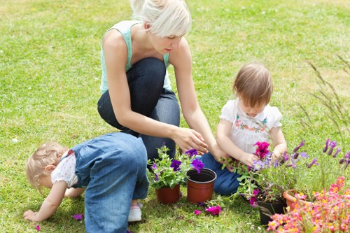A woman with short blonde hair engaged in gardening activities with two young children in a well-maintained backyard garden. The woman, dressed in a light blue sleeveless top and dark jeans, is kneeling on the grass and planting or tending to flowering plants in small pots. One child, wearing a denim overall, is bent over close to the ground, possibly picking up fallen flowers or inspecting the soil, while the other child, in a white top with pink floral patterns, is sitting nearby, tending to a cluster of purple and pink flowering plants. The garden features a lush green lawn extending across the compacted soil areas, bordered by flower beds containing vibrant perennials. To the right side, there are lush shrubs and small trees, suggesting a mature landscaped garden designed for outdoor leisure and family gardening activities. The scene is outdoors during daytime with natural light, under cloudy or partly sunny weather conditions, creating a calm and natural outdoor environment. This image reflects typical gardening and outdoor maintenance activities suitable for residential properties in Barkingside or surrounding areas, highlighting the landscape features that Gardening Services Barkingside might service.