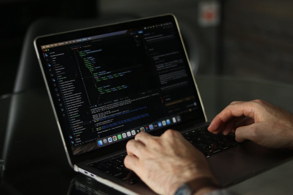 A person using a laptop on a dark surface, with laptop screen displaying lines of colorful code, indicating programming or web development work. The individual's hands are positioned over the keyboard